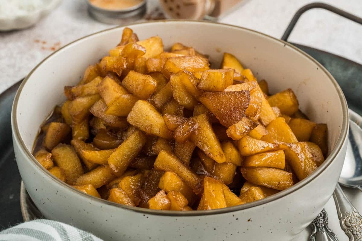 A white bowl filled with cooked, diced apples coated in brown cinnamon sauce, sitting on a tray with a spoon nearby.