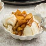 A bowl of vanilla ice cream topped with spiced cooked apple chunks, served with a spoon on a light-colored surface.