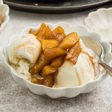 A bowl of vanilla ice cream topped with spiced cooked apple chunks, served with a spoon on a light-colored surface.