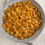 Chopped apples cooked with spices in a large, silver frying pan on a light-colored surface.