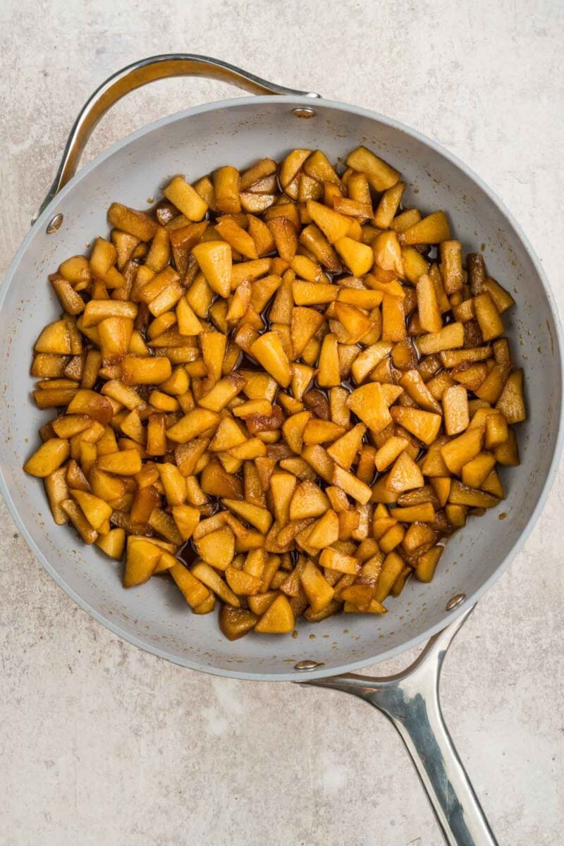 Chopped apples cooked with spices in a large, silver frying pan on a light-colored surface.