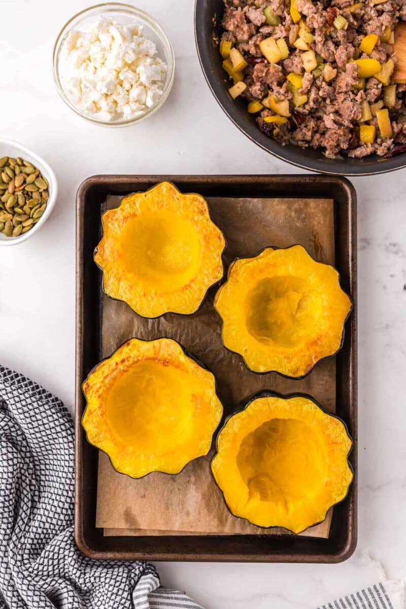 Four roasted acorn squash halves on a baking sheet, with cooked ground meat and diced vegetables in a pan, and bowls of feta cheese and pepitas nearby.