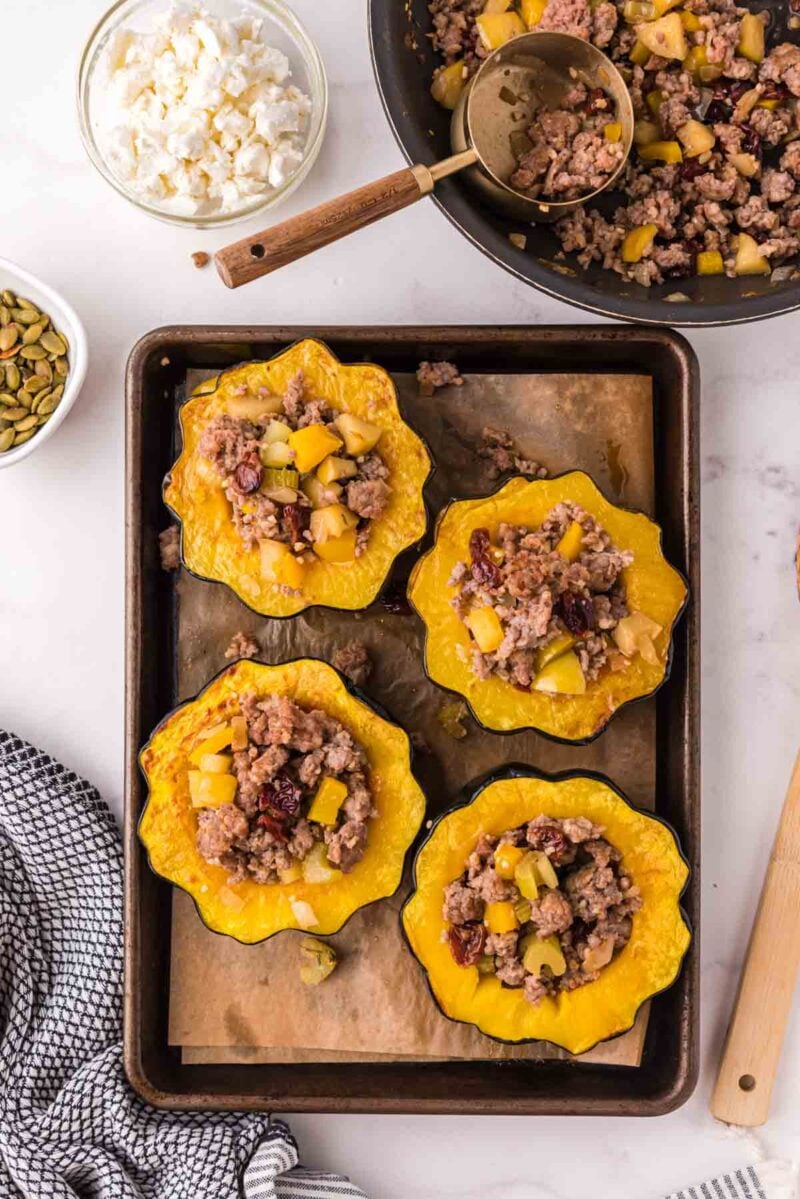Four halved acorn squashes filled with a ground meat, apple, and cranberry mixture on a baking tray, with bowls of cheese and pepitas nearby.