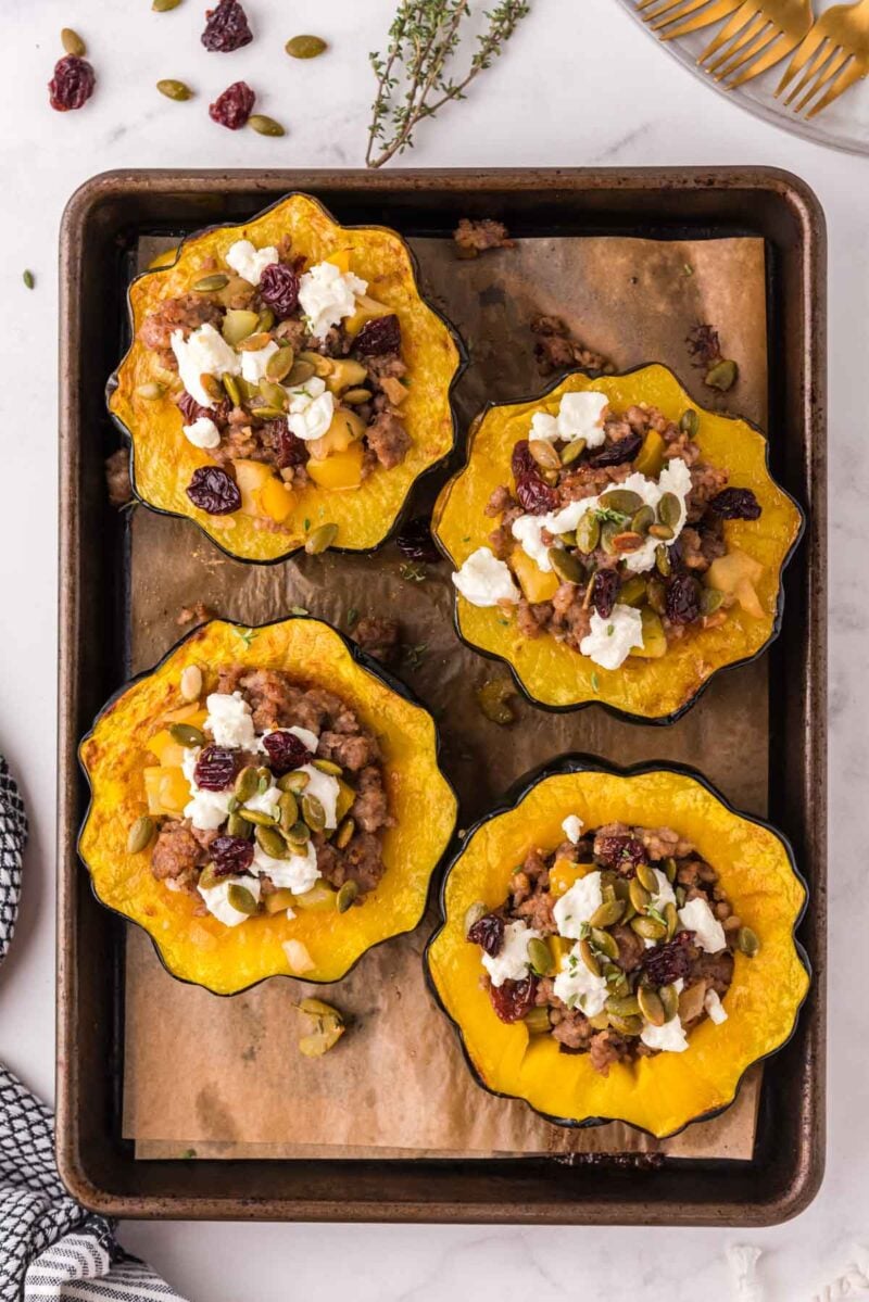 Four baked acorn squash halves filled with a mixture of ground meat, cheese, seeds, and dried fruit on a parchment-lined baking sheet.