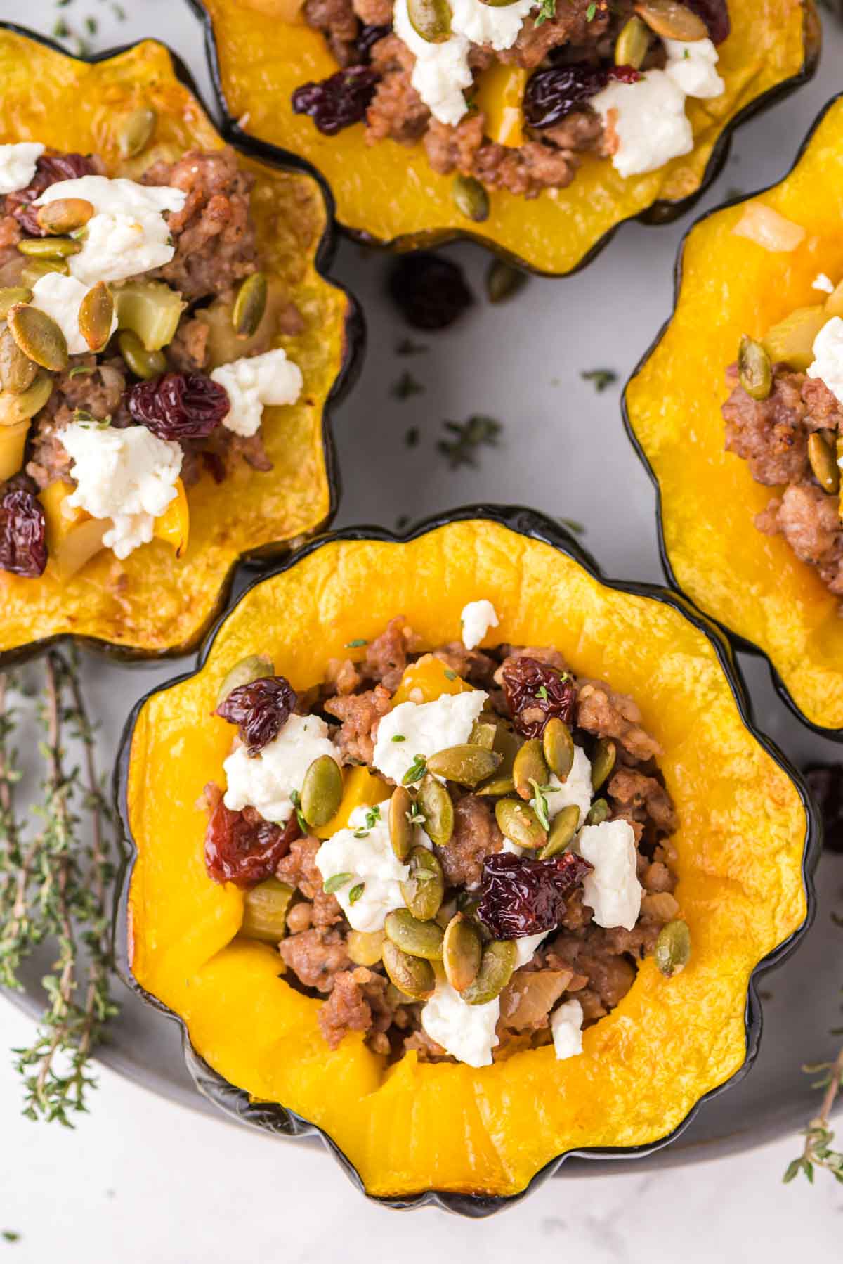 Four halved acorn squashes filled with a stuffing of ground meat, feta cheese, pumpkin seeds, and dried cranberries, arranged on a plate with fresh herbs nearby.