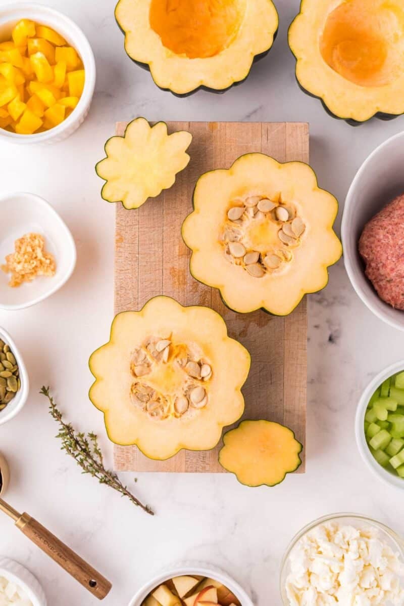 Two halved acorn squashes with seeds on a cutting board, surrounded by bowls of diced vegetables, ground meat, herbs, and cheese on a white surface.