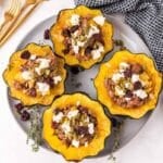 Four stuffed acorn squash halves filled with a mixture of meat, cheese, seeds, and dried cranberries, arranged on a gray plate with herbs, next to gold utensils and a black and white towel.