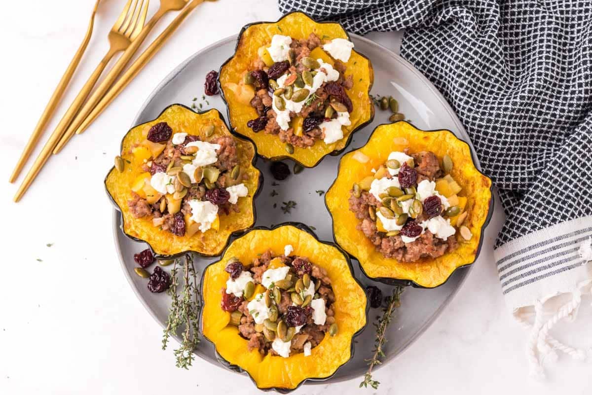 Four stuffed acorn squash halves filled with a mixture of meat, cheese, seeds, and dried cranberries, arranged on a gray plate with herbs, next to gold utensils and a black and white towel.