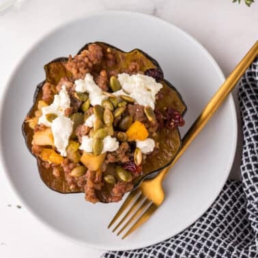 Stuffed acorn squash filled with ground meat, cheese, seeds, and vegetables on a white plate with a gold fork and knife, next to a black and white checkered napkin.