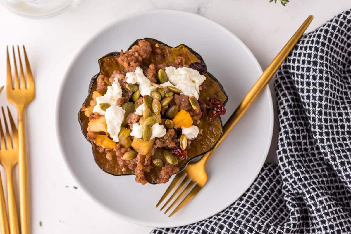 Stuffed acorn squash filled with ground meat, cheese, seeds, and vegetables on a white plate with a gold fork and knife, next to a black and white checkered napkin.