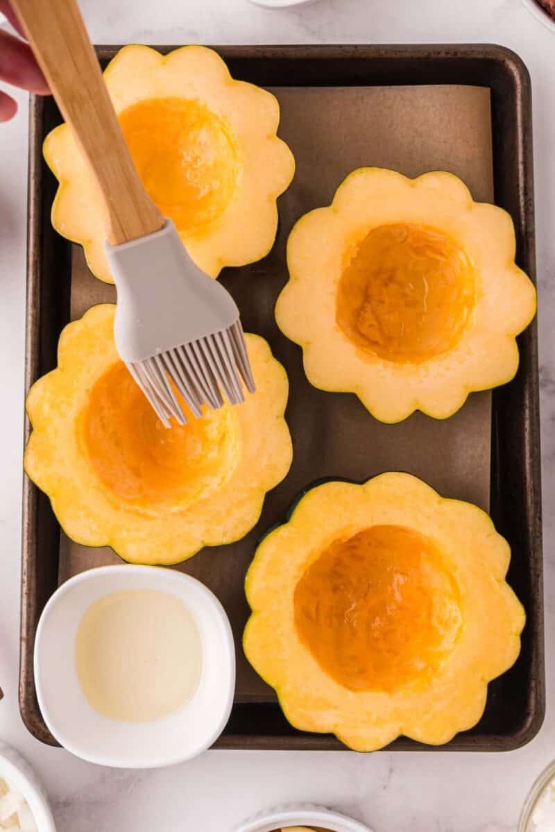 Four halved acorn squash on a baking tray are being brushed with oil using a silicone brush. Small bowls with ingredients are nearby.