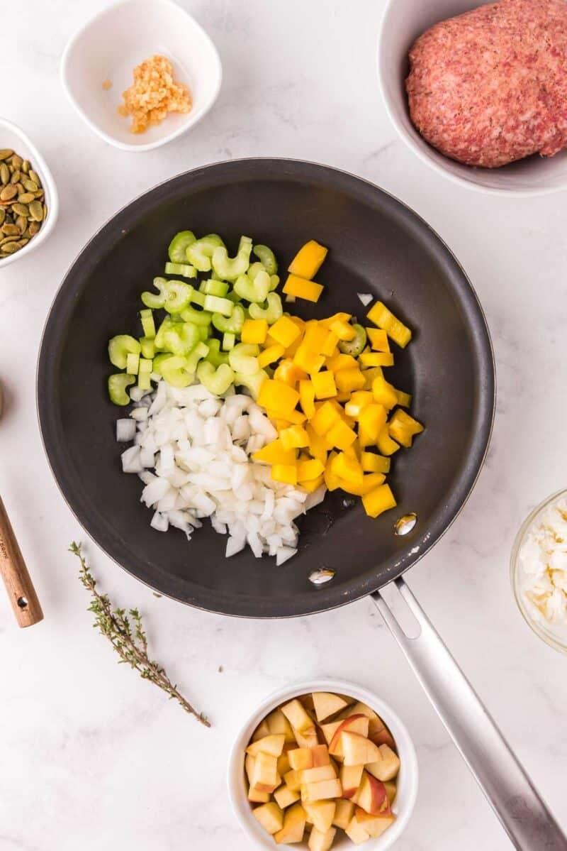 A frying pan with chopped celery, yellow bell pepper, and onion, surrounded by bowls of diced apple, ground meat, garlic, seeds, and fresh thyme on a white surface.