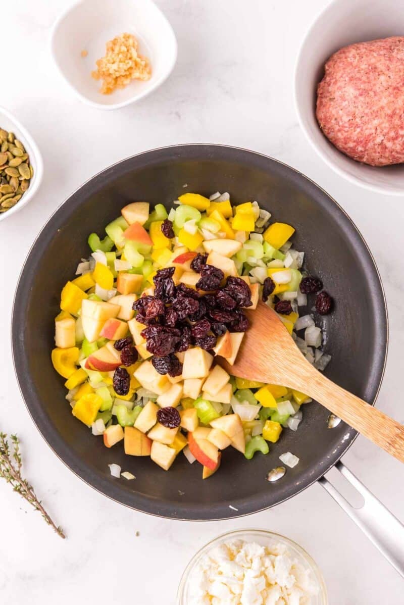 A skillet with chopped onions, celery, apples, yellow bell pepper, and dried cranberries being stirred with a wooden spoon; raw ground meat and other ingredients nearby.