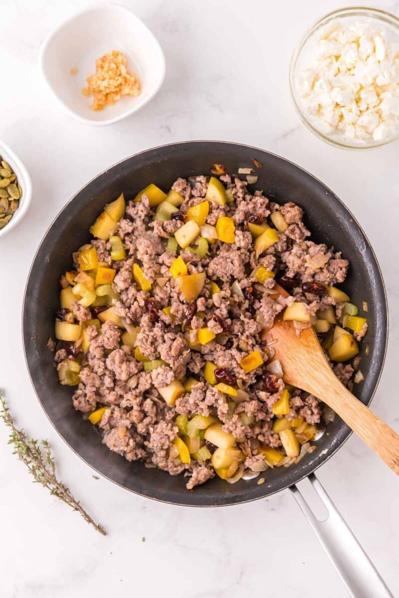 A skillet filled with cooked ground meat, diced yellow vegetables, and herbs, with a wooden spoon. Nearby are bowls of crumbled cheese, seeds, and minced garlic, plus a sprig of thyme.