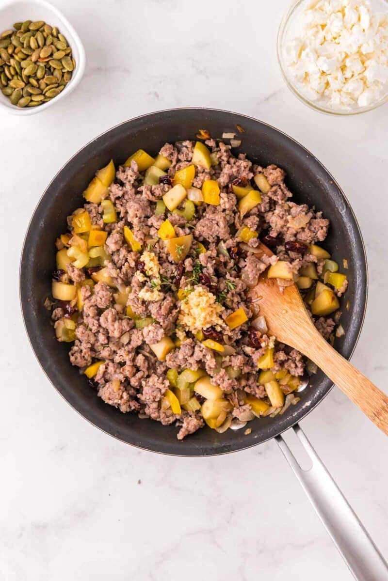 A skillet filled with cooked ground meat, diced vegetables, and seasonings is stirred with a wooden spoon. Nearby are small bowls of pumpkin seeds and crumbled cheese.