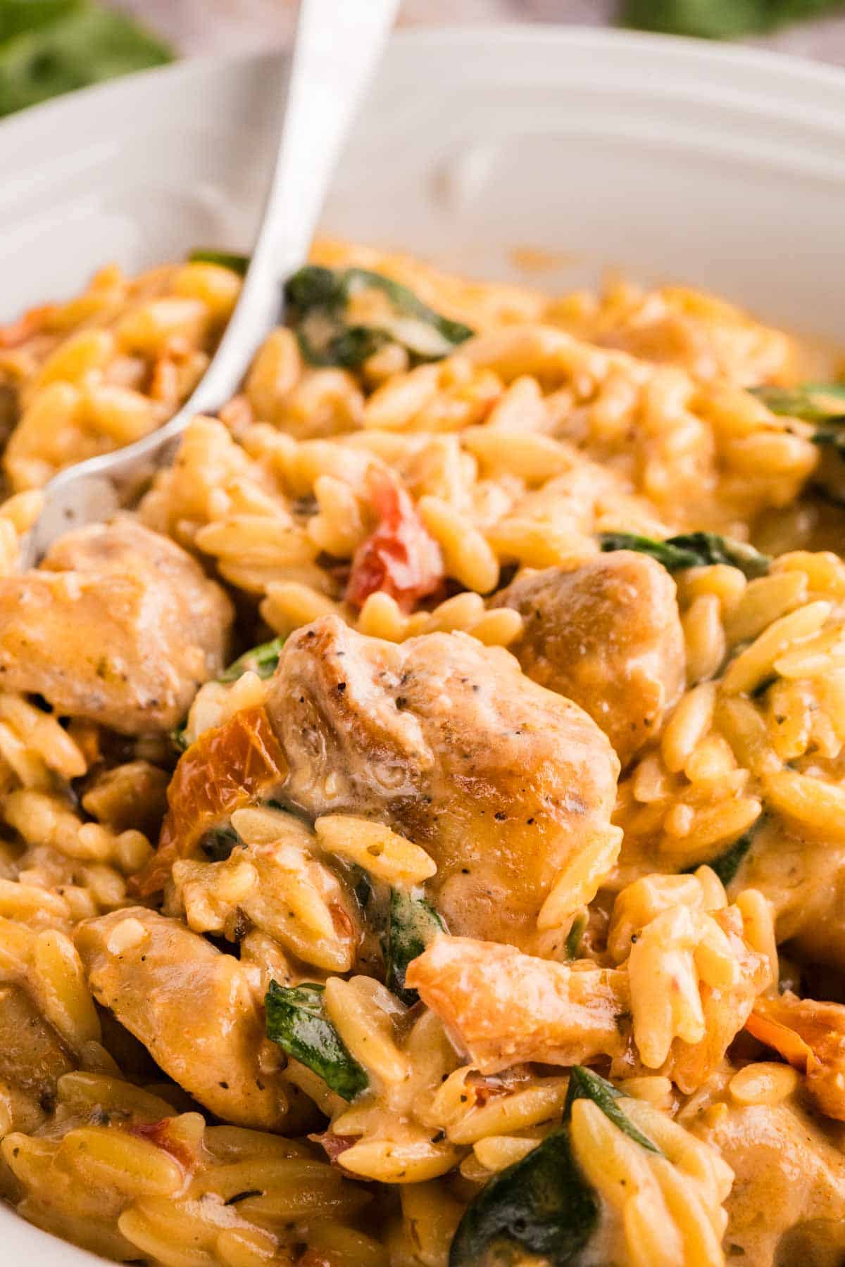 Close-up of creamy orzo pasta with pieces of cooked chicken, spinach, and tomatoes, served in a white bowl with a fork.