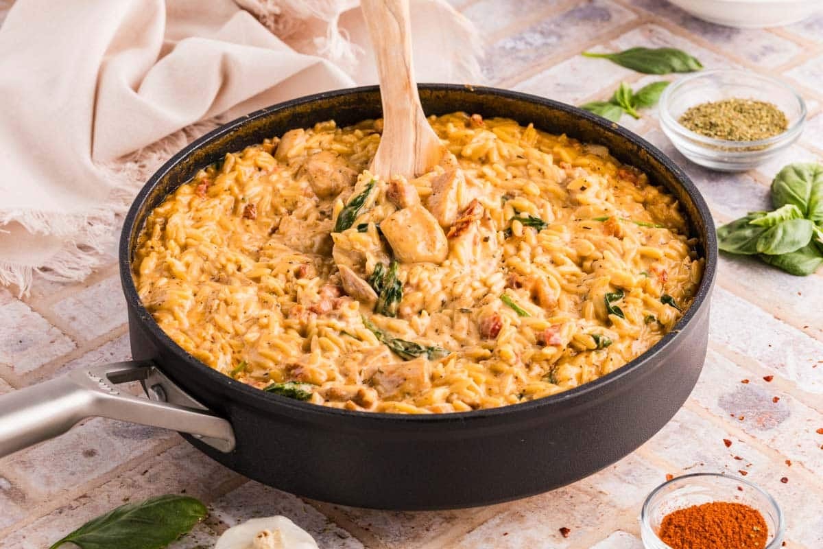 A skillet filled with creamy orzo pasta with chicken, spinach, and herbs, being stirred with a wooden spoon.