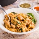 A white bowl of creamy orzo pasta with chicken, spinach, and herbs, with a spoon inside; spice bowls and basil leaves are in the background.
