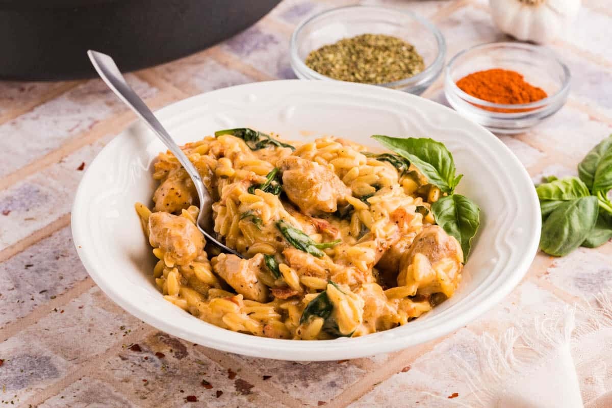 A white bowl of creamy orzo pasta with chicken, spinach, and herbs, with a spoon inside; spice bowls and basil leaves are in the background.