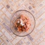A glass bowl containing raw, chopped chicken pieces with salt and pepper on a light-colored tile surface.