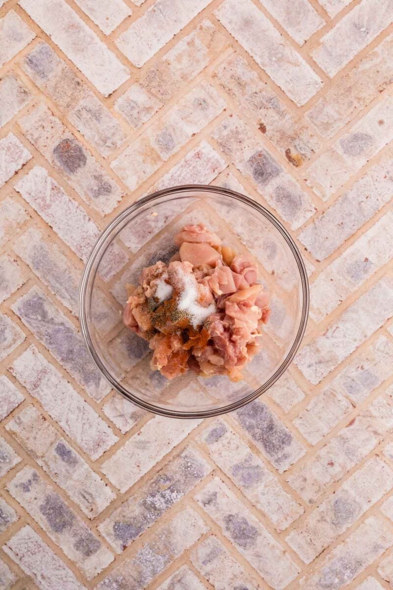 A glass bowl containing raw, chopped chicken pieces with salt and pepper on a light-colored tile surface.