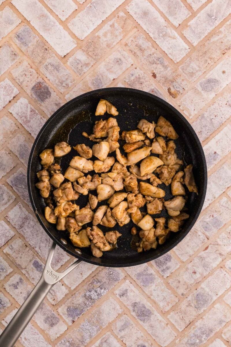A frying pan containing cooked, seasoned chicken pieces sits on a light-colored, patterned tile surface.