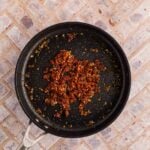 A nonstick frying pan with cooked, finely chopped sun-dried tomatoes and herbs, seen from above on a light brick-patterned surface.