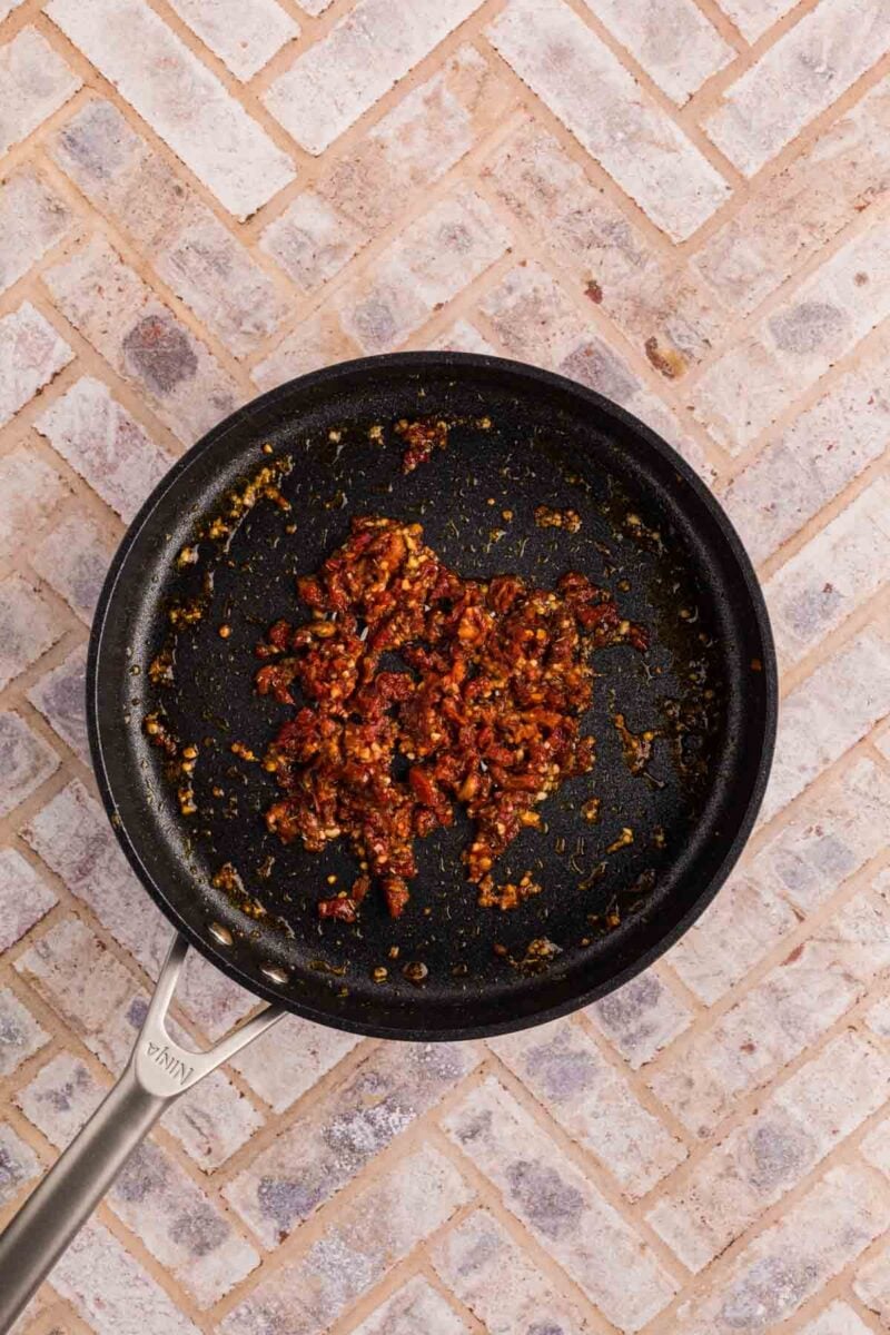 A nonstick frying pan with cooked, finely chopped sun-dried tomatoes and herbs, seen from above on a light brick-patterned surface.