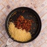 A black skillet on a tiled surface contains uncooked orzo pasta and a cooked tomato-based mixture.