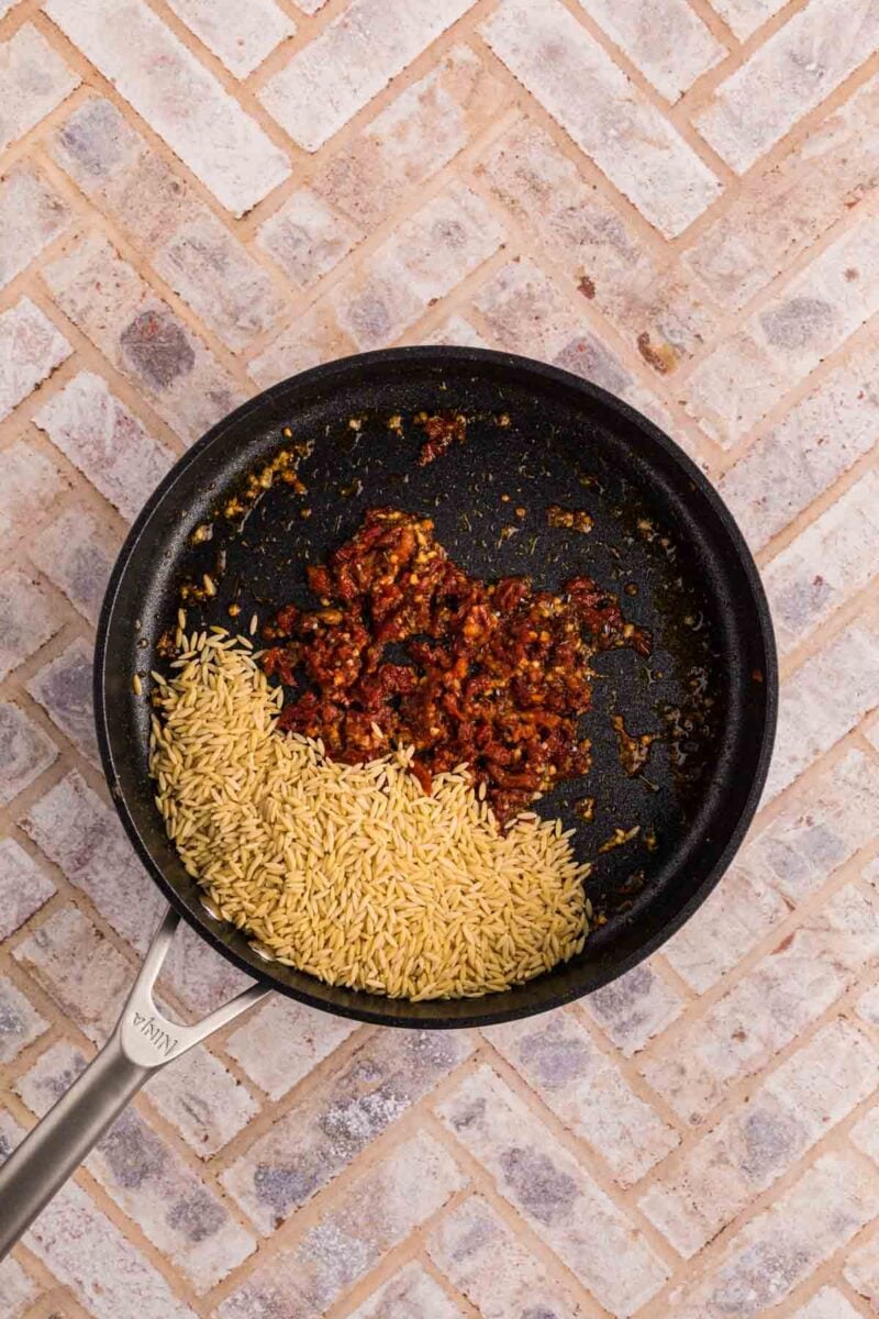 A black skillet on a tiled surface contains uncooked orzo pasta and a cooked tomato-based mixture.