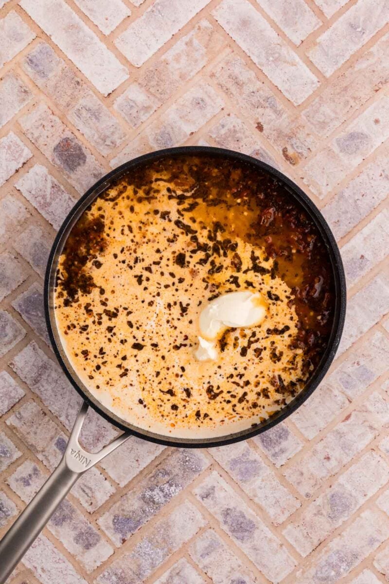 A skillet with a creamy sauce and browned bits, topped with a dollop of butter, sits on a light tile surface.
