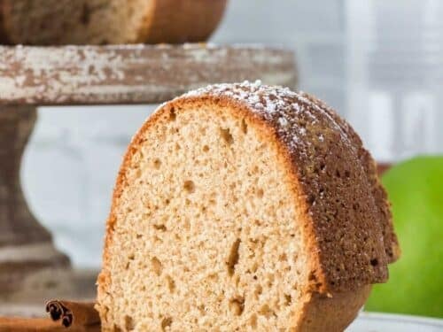 A slice of cinnamon bundt cake sits on a plate with two cinnamon sticks beside it; the cake is lightly dusted with powdered sugar.