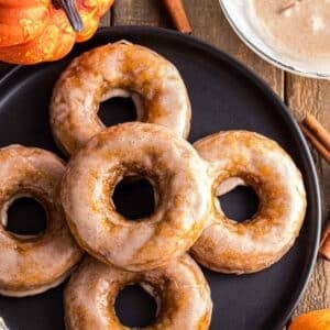 Five glazed donuts are arranged on a black plate, with a bowl of glaze, cinnamon sticks, and a small pumpkin nearby on a wooden table.