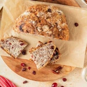 A round loaf of rustic bread with visible nuts and cranberries, partially sliced, sits on parchment paper atop a wooden board. Several bread slices and loose cranberries and pecans are nearby.