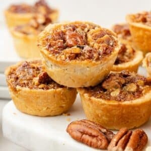 Three pecan tarts stacked on a white surface, with additional tarts and whole pecans scattered around them.