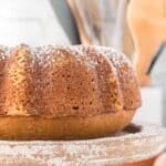 A bundt cake dusted with powdered sugar sits on a rustic cake stand, with kitchen utensils blurred in the background.