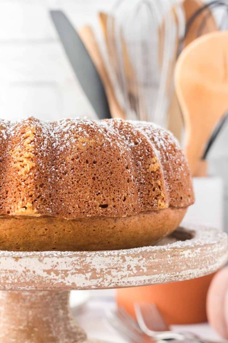 A bundt cake dusted with powdered sugar sits on a rustic cake stand, with kitchen utensils blurred in the background.