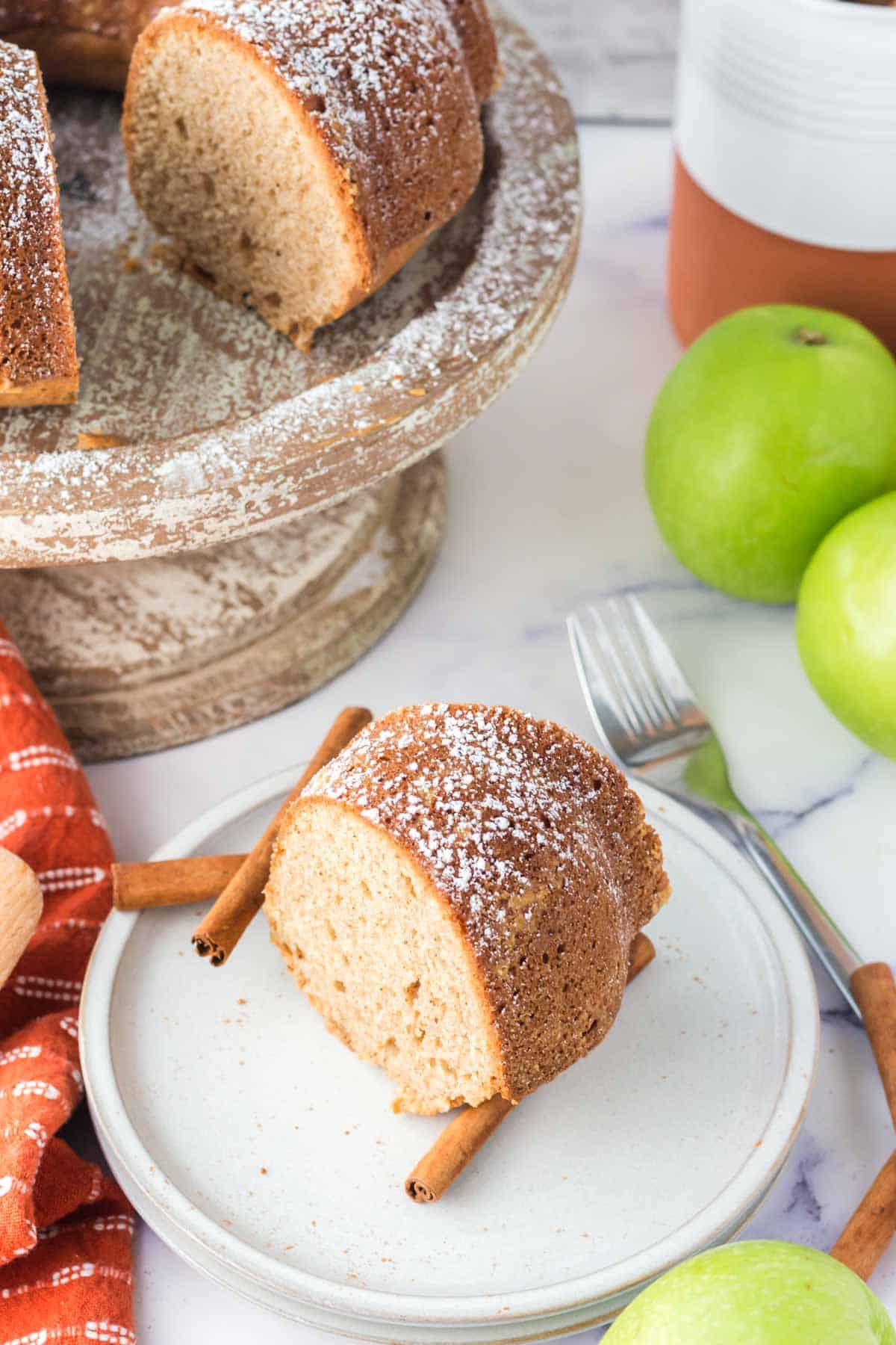 A slice of bundt cake dusted with powdered sugar on a plate with cinnamon sticks, next to whole green apples and a fork, with more cake on a stand in the background.