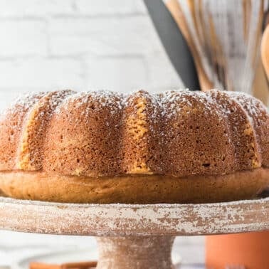 A round bundt cake with a light dusting of powdered sugar sits on a rustic cake stand; kitchen utensils are visible in the background.