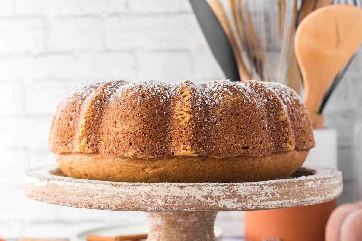 A round bundt cake with a light dusting of powdered sugar sits on a rustic cake stand; kitchen utensils are visible in the background.