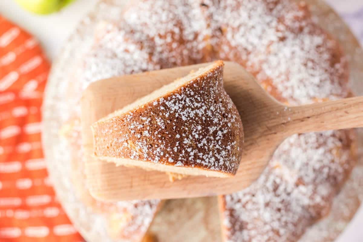 A wooden spatula lifts a powdered sugar-dusted slice of round cake from the whole cake, viewed from above.