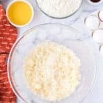 A glass bowl of creamed butter and sugar sits on a marble counter, surrounded by flour, melted butter, eggs, vanilla, yogurt, cinnamon, and a red dish towel.