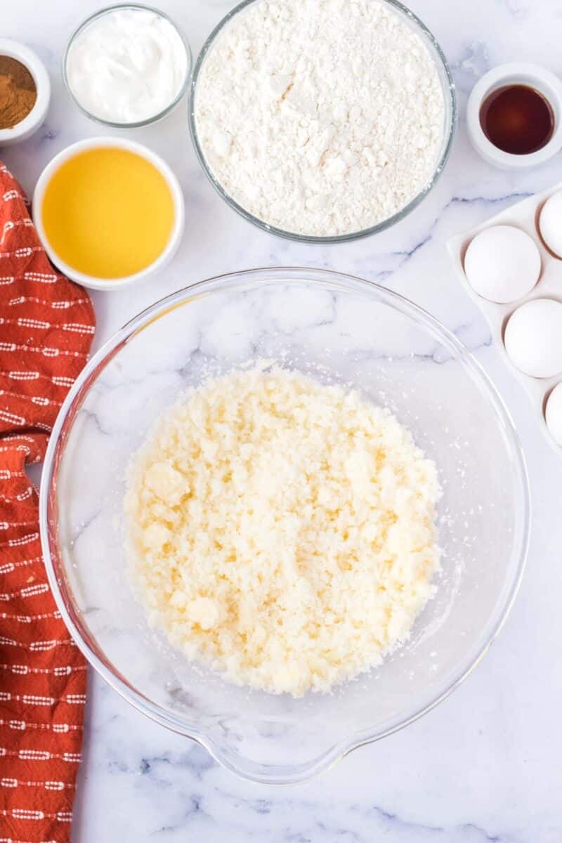 A glass bowl of creamed butter and sugar sits on a marble counter, surrounded by flour, melted butter, eggs, vanilla, yogurt, cinnamon, and a red dish towel.
