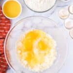 A glass bowl with four cracked eggs and creamed mixture, surrounded by flour, orange juice, sour cream, vanilla, and empty eggshells on a marble counter.
