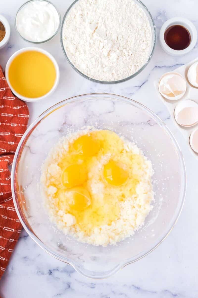 A glass bowl with four cracked eggs and creamed mixture, surrounded by flour, orange juice, sour cream, vanilla, and empty eggshells on a marble counter.