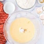 A glass bowl with whisked wet ingredients, a bowl of flour, cracked eggshells, and small bowls of baking ingredients on a marble countertop.