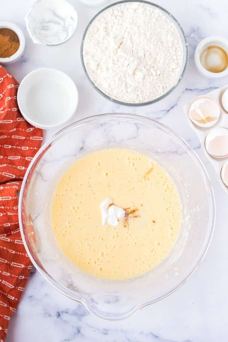 A glass bowl with whisked wet ingredients, a bowl of flour, cracked eggshells, and small bowls of baking ingredients on a marble countertop.