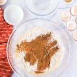 Large glass bowl with flour and cinnamon mixture, surrounded by empty bowls, eggshells, a red kitchen towel, and ingredients on a marble countertop.