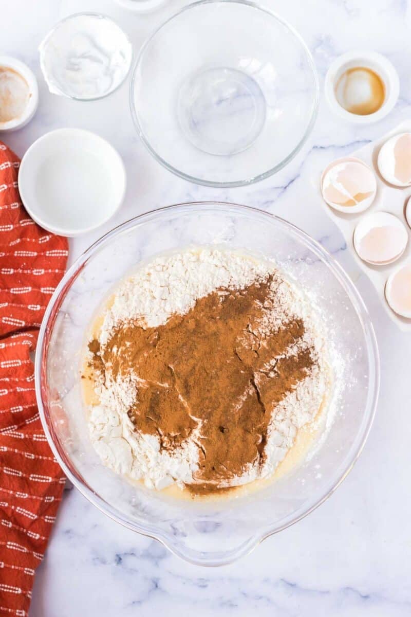 Large glass bowl with flour and cinnamon mixture, surrounded by empty bowls, eggshells, a red kitchen towel, and ingredients on a marble countertop.