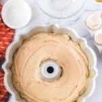Bundt pan filled with unbaked cake batter on a marble countertop, surrounded by baking ingredients, a mixing bowl, cracked eggshells, and a red dish towel.